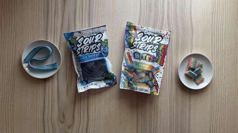 A bag of Sour Strips Blue Raspberry and Sour Strip Bites Rainbow on a table beside two small bowls filled with the candy