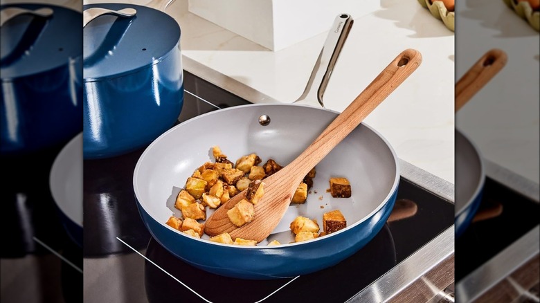 Blue ceramic pan on a stovetop with potato cubes sautéed with a wooden spoon.