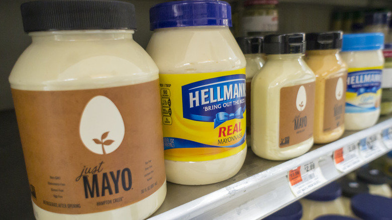 Several different brands of jarred mayonnaise sitting on a grocery store shelf.