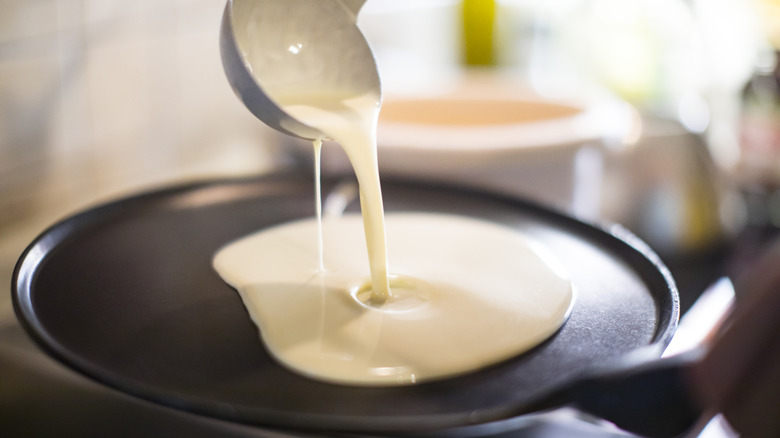 A ladle pours pancake batter onto a hot frying pan.