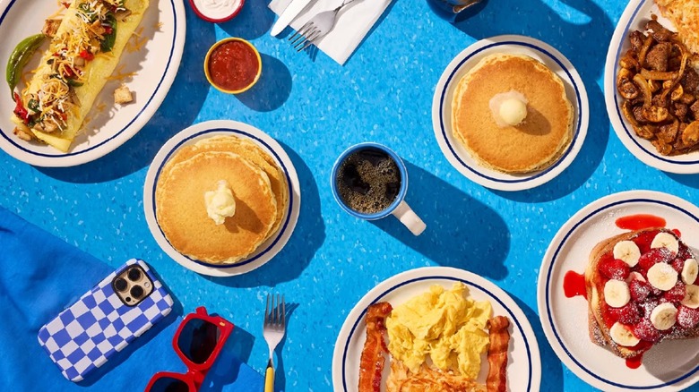 Breakfast dishes from IHOP on white plates sitting on blue surface, including strawberry banana French toast, pancakes with butter, eggs with bacon, and omelet