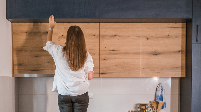 A woman motioning to open brown wooden cabinets in a kitchen.