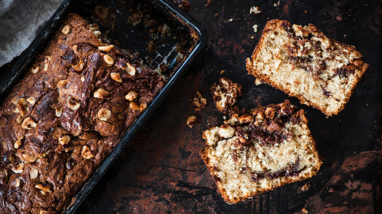 A pan containing a sliced loaf topped with hazelnuts sitting alongside two slices banana bread with dark swirls