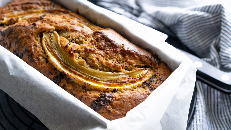 A loaf of banana bread resting in a parchment paper lined pan on top of a blue and white striped cloth