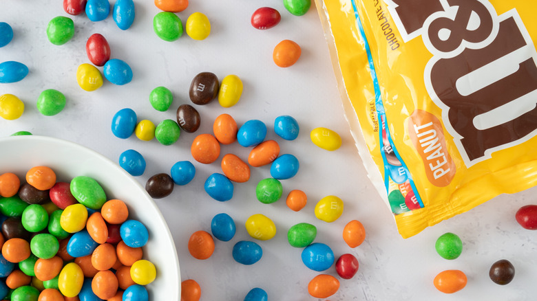 An open bag of peanut M&Ms spilling out onto a surface next to a bowl of more M&Ms.