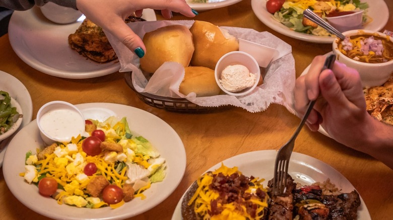 Bowl of salad, plate with steak and loaded potato, plate with seafood and chili, and hands reaching into basket of buns
