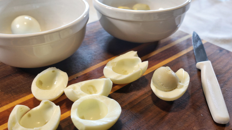 halved boiled eggs on a wooden cutting board with a knife, a bowl of egg yolks, and a bowl of egg whites