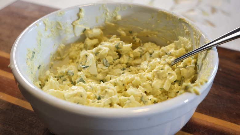 a bowl of egg salad with a spoon in it on a wooden cutting board