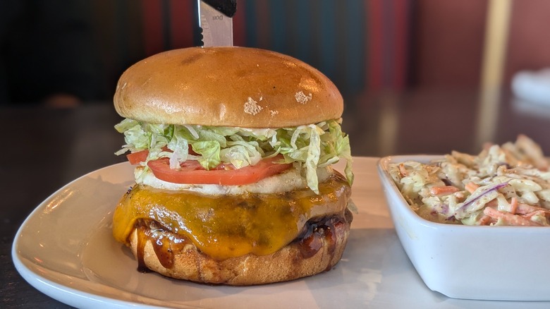 Close-up of burger with cheddar, lettuce, tomato, and pineapple and side of coleslaw on white plate