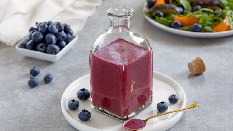 A small jug of blueberry vinaigrette on a plate with a spoon next to a bowl of blueberries and a salad