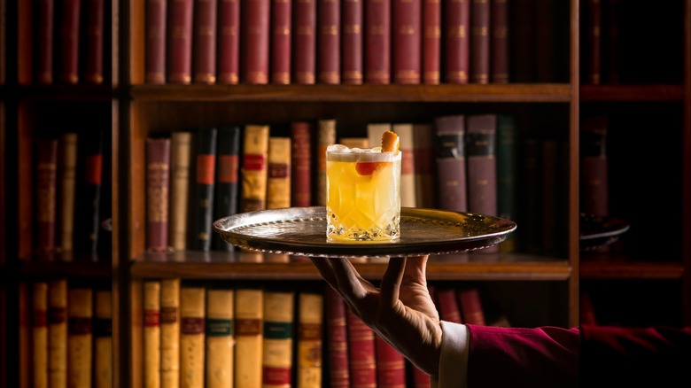A hand holding a platter with a cocktail in front of a bookcase