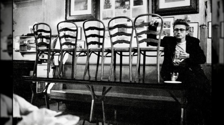 James Dean holding a drink and leaning on chairs that are on top of a table