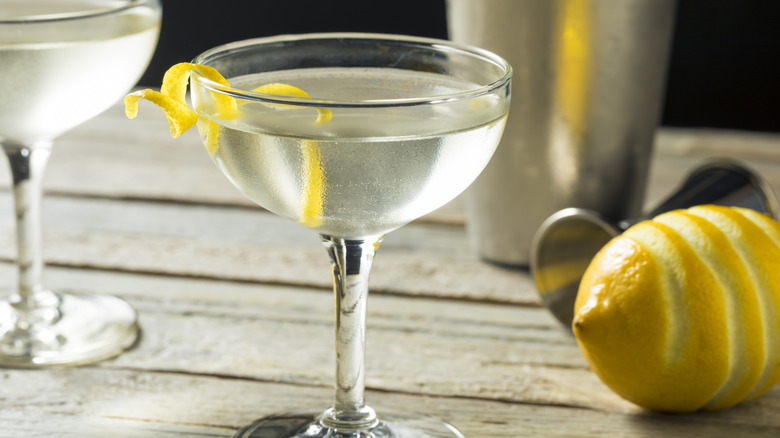 dry vodka martini with a twist on a table with another glass, a metal cocktail shaker, and a lemon.