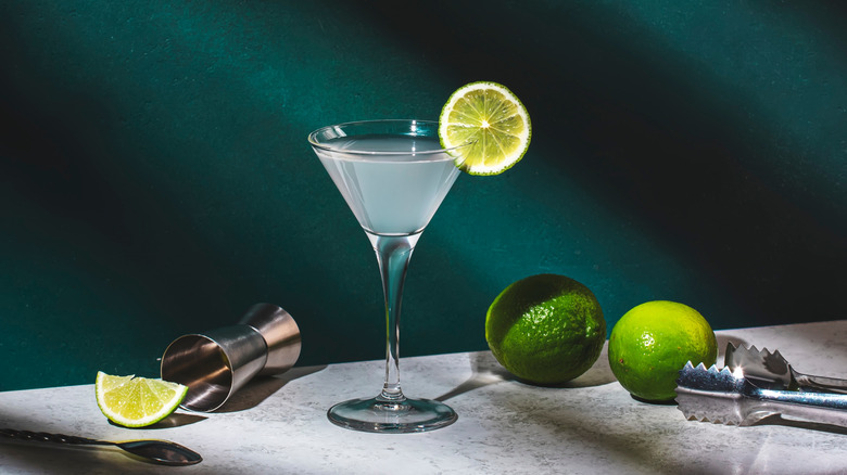 gimlet cocktail surrounded by limes and cocktail equipment against a dark green backdrop.