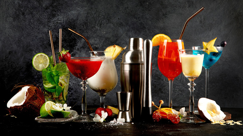 a lineup of cocktails of various colors, shapes, and sizes with a metal cocktail shaker in the middle and ingredients on the table.