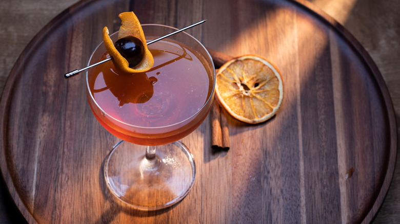 manhattan cocktail with dark cherry and orange garnish on a wooden cutting board.