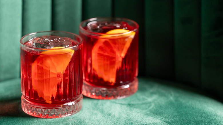 two classic red negronis with orange slices in them against a jewel toned green velvet backdrop.