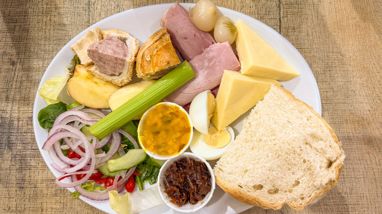 a ploughman's lunch featuring cheese, meats, veggies, eggs and bread on a white plate