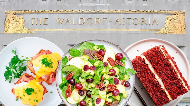 Three white plates with eggs Benedict, Waldorf salad, and red velvet cake in front of Waldorf-Astoria hotel building