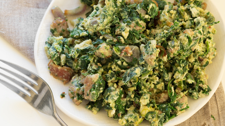 A plate of Joe's Special featuring spinach and scrambled eggs with visible bits of mushroom and ground meat. There is a fork to the left of the plate