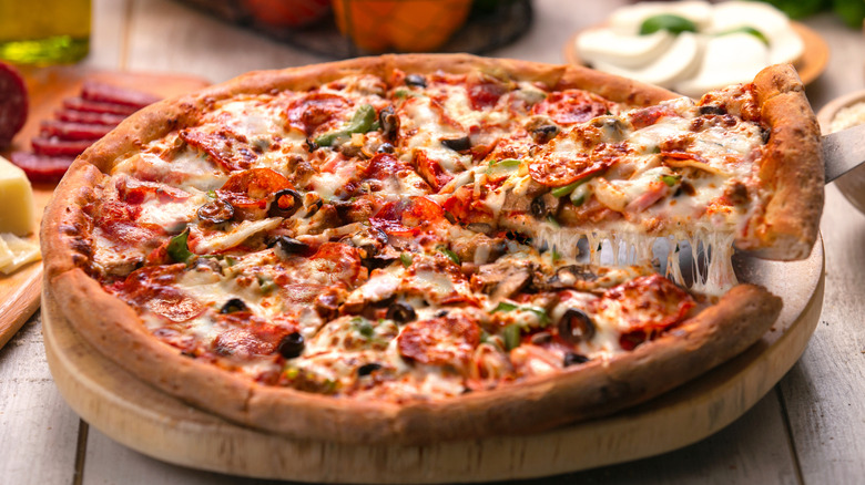 Supreme pizza cut into slices on wooden board with serving knife lifting up a single slice