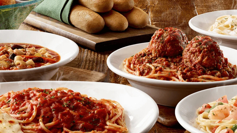 Several bowls of pasta sitting on a counter top with breadsticks nearby