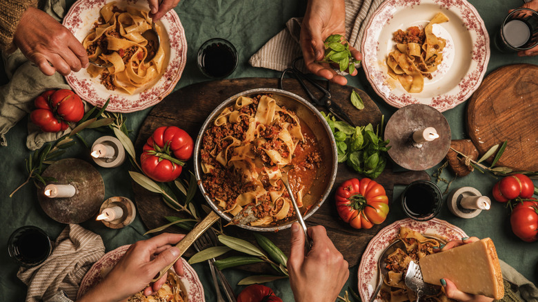 An overhead shot of a full dinner table with a pot of bolognese pasta at the center, herbs and tomatoes on the table, and hands and plates of happy diners