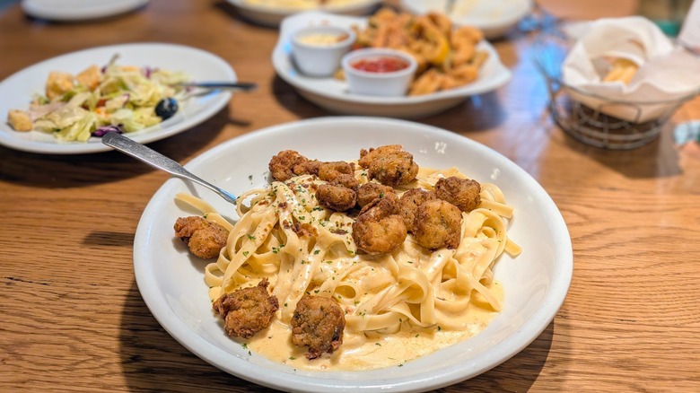 A bowl of fettucini with Alfredo sauce and crispy shrimp with a plate of salad and a fried appetizer in the background on a wood table