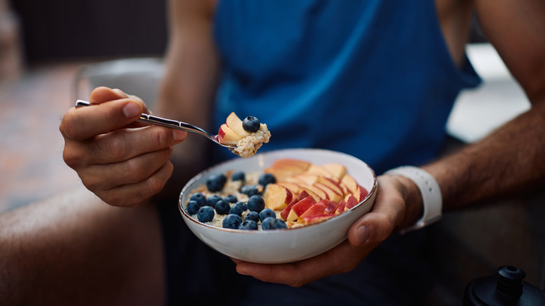 Person dressed in athletic wear holding spoon and white bowl of oatmeal topped with fresh fruit