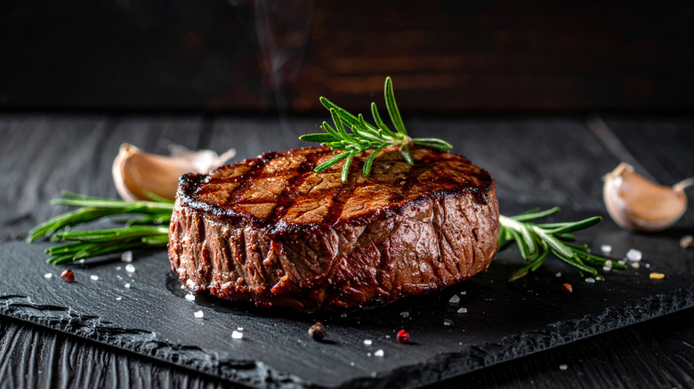 A grilled steak on a stone platter with rosemary, garlic, salt, and peppercorns around it