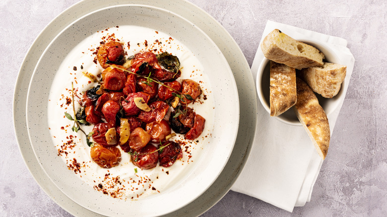 A creamy dip topped with roasted tomatoes and herbs next to a ramekin of sliced baguette