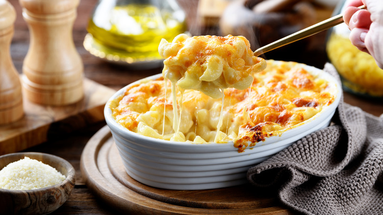 Someone lifting a spoonful of creamy, cheesy baked mac and cheese out of a white bowl that is sitting on a wooden trivet