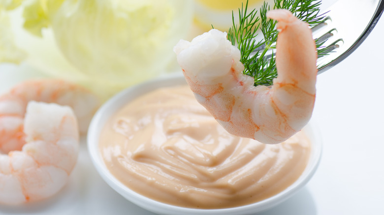 A cooked, peeled shrimp on a fork with fresh dill about to be dipped into a small dish of creamy pink sauce.