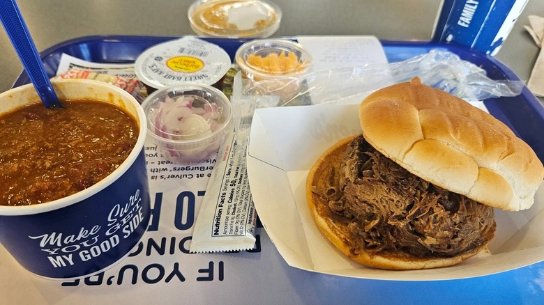 close up of a Culver's beef pot roast sandwich, blue cup of chili, and various condiments and toppings on blue dining tray.