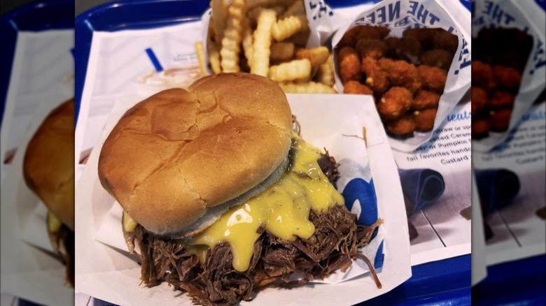 A Culver's pot roast sandwich with cheese sauce sitting on tray next to paper sleeves of French fries and cheese curds.