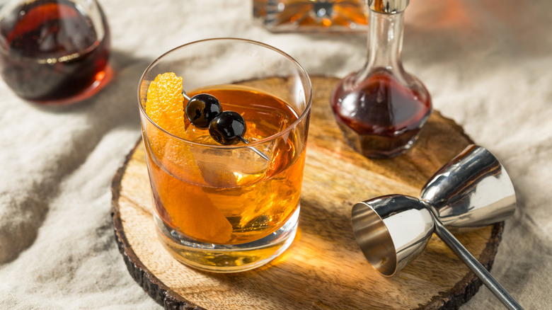 Old Fashioned cocktail sitting on a wooden platter beside a jigger and a small glass bottle of maple syrup