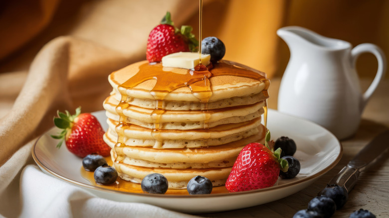 Stack of pancakes with berries, butter, and maple syrup getting drizzled on with a white pitcher in the background.