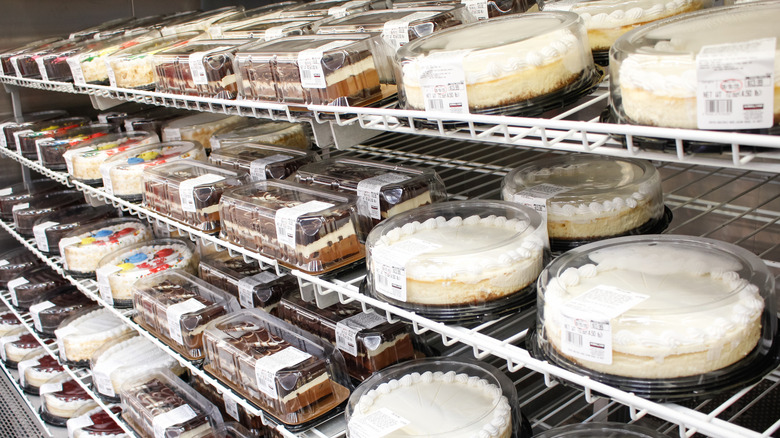 Various cakes include cheesecakes, bar cakes, and birthday cakes on display shelves in Costco store