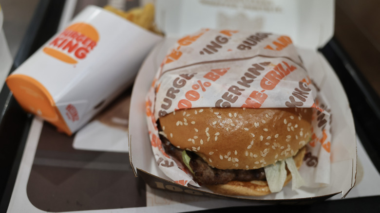 A burger king burger sits in a container on a plastic tray next to a box of fries