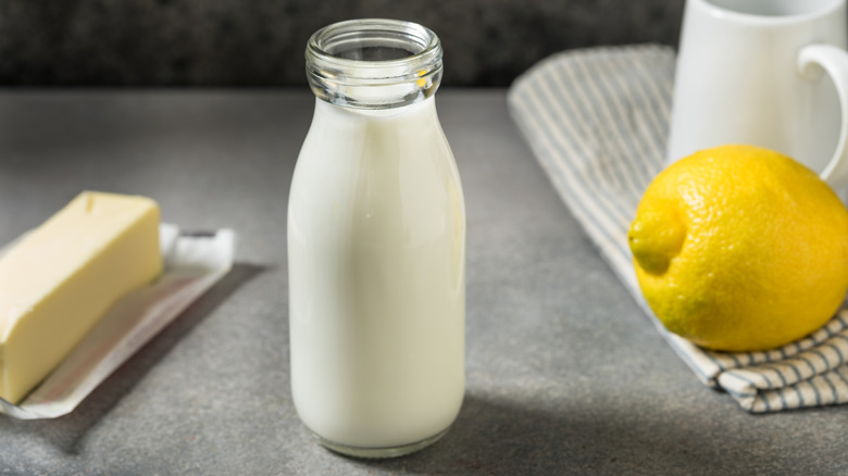 A jar of buttermilk on a grey counter and backdrop with a stick of butter, linen napkin, lemon, and ceramic cup in the background
