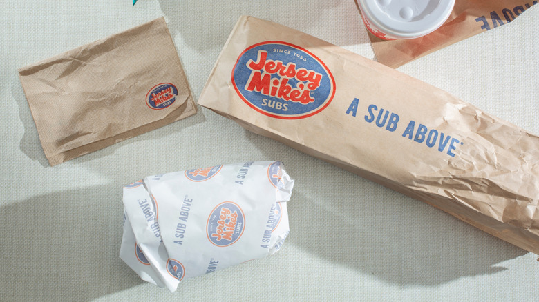 A wrapped Jersey Mike's sub, the paper bag, napkins, and a drink cup on a white table