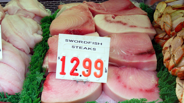 Raw swordfish steak on display with other seafood and a sign indicating it's selling for $12.99 a pound