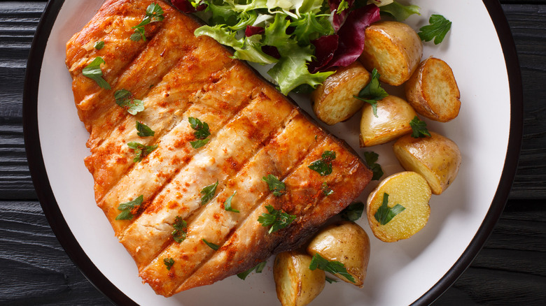 A golden crusted swordfish steak on a plate with some baby potatoes and spring mix salad