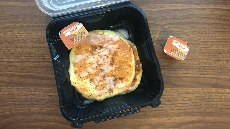 Denny's Cinnamon Roll pancakes in a plastic to-go container on top of a wooden table with two containers of syrup
