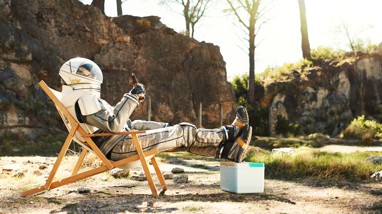 a person in a space suit holding a beer and reclining in a chair with their feet up on a small cooler