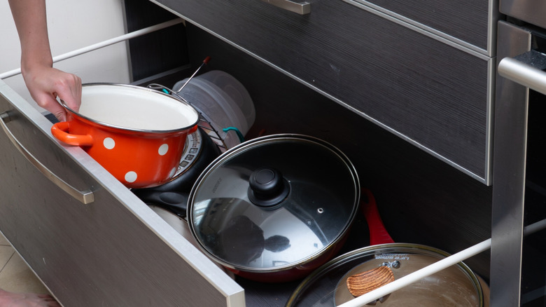 A boy holding a frying pan with his face unseen reaches into a deep kitchen drawer that is cluttered with pots and pans