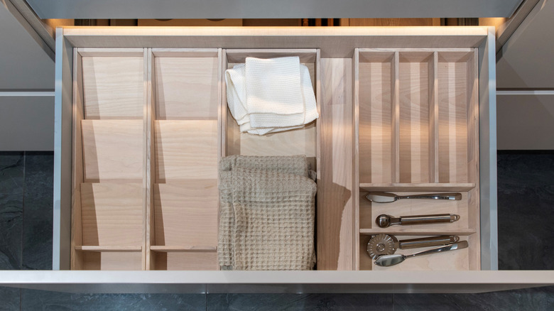 A wooden stackable organizer in a kitchen drawer, containing several utensils and kitchen towels in their own distinct sections