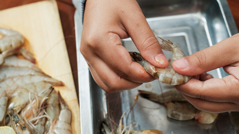 two hands peeling raw shrimp over a metal container with more shrimp to the right.