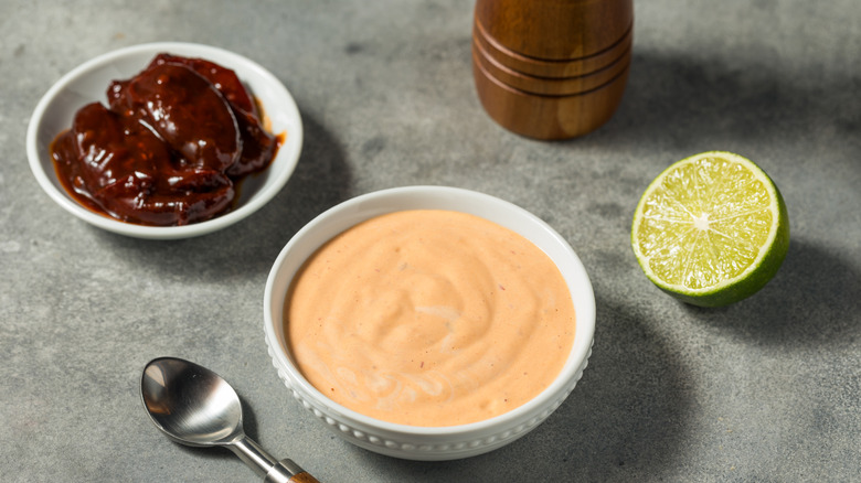 A bowl of spicy sauce sits on a table next to half a lime and a bowl of chipotle peppers