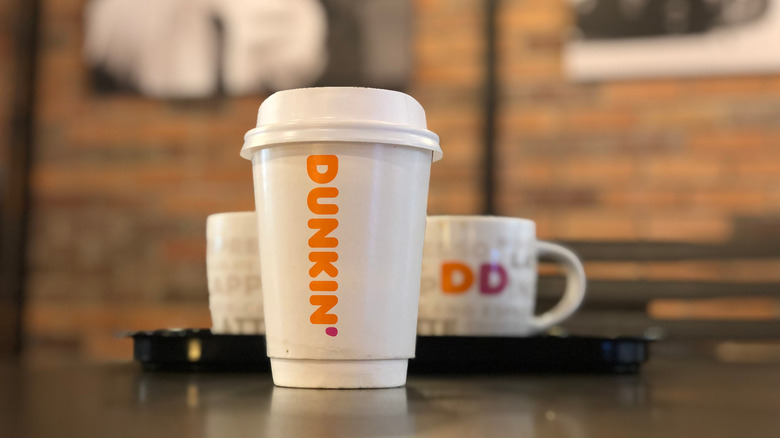 A hot Dunkin' coffee in a to-go cup with two mugs on a tray seen in the background